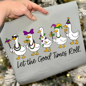 May include: A light grey fabric featuring a cartoon design of five white ducks in Mardi Gras costumes. Each duck is adorned with various accessories, such as sunglasses, masks, and hats. The text "Let the Good Times Roll" is printed in black below.