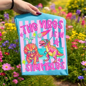 Peut inclure: T-shirt bleu clair avec l'inscription rose "THE VIBES IS IN SHAMBLES". Le motif pr&eacute;sente des dinosaures de dessins anim&eacute;s, une m&eacute;t&eacute;orite et des &eacute;toiles. Les dinosaures portent des lunettes de soleil et un &eacute;quipement de plong&eacute;e.