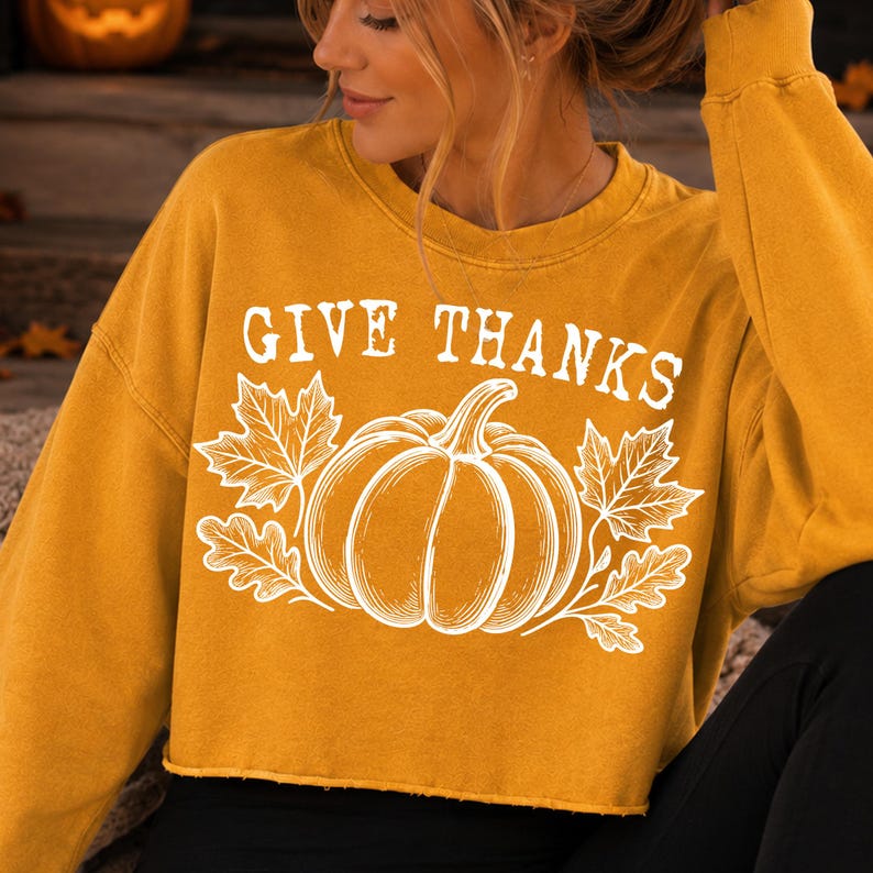 Puede incluir: Una sudadera corta de manga larga en color amarillo mostaza con un estampado gr&aacute;fico blanco de una calabaza rodeada de hojas de arce y roble. El texto "GIVE THANKS" aparece en una fuente serif blanca y negrita sobre la calabaza.