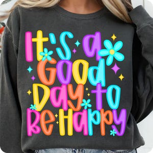 K&ouml;nnte beinhalten: Ein schwarzer Pullover mit dem Text "It's a good day to be happy" in bunten Buchstaben und Blumen. Der Text ist in einer Schriftart im Retro-Stil.