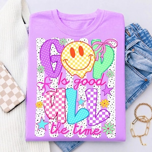 Op de afbeelding: Een lichtpaars t-shirt met een grafische print met de tekst "God is good all the time". Het ontwerp bevat een geel geblokt smileygezicht, kleurrijke geblokte letters, roze strikken, kleine sterretjes en bloemen op een zwarte achtergrond met stippen.