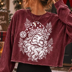 K&ouml;nnte beinhalten: Ein kastanienbraunes, lang&auml;rmeliges bauchfreies Sweatshirt mit einem wei&szlig;en Grafikdruck eines zwinkernden Weihnachtsmanns, der eine M&uuml;tze mit Leoparden- und Punktemuster tr&auml;gt, umgeben von Tannennadeln und Stechpalmenbeeren auf der Vorderseite.