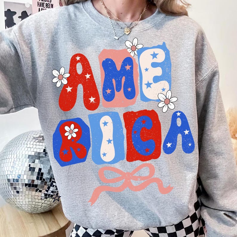 May include: A grey sweatshirt featuring the word "AMERICA" in red, white, and blue block letters. The letters are decorated with stars and flowers. A pink bow is at the bottom of the design.