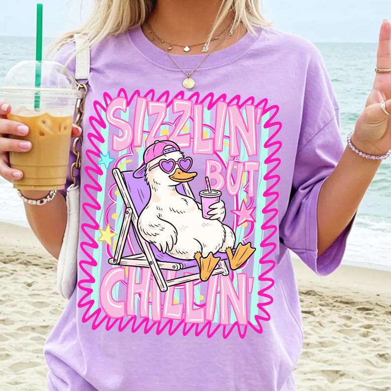Op de afbeelding: Een lila T-shirt met een afbeelding van een eend met een zonnebril en een hoed, ontspannen in een strandstoel. De tekst op de shirt luidt "Sizzlin' But Chillin'" in een roze en paars ontwerp. Het shirt heeft korte mouwen.