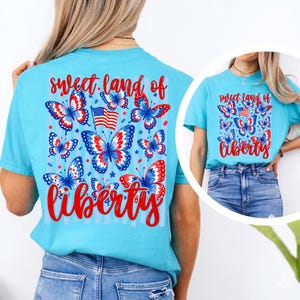 May include: A light blue t-shirt featuring a graphic print of red, white, and blue butterflies, a small American flag, and the red text "sweet land of liberty". The design is displayed on the back of the shirt and also on the front in a circular inset.