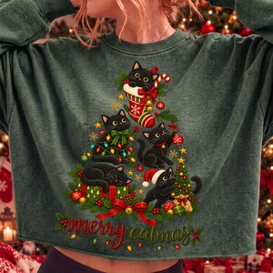 K&ouml;nnte beinhalten: Ein dunkelgr&uuml;nes Langarm-Sweatshirt mit einer Grafik von vier schwarzen Katzen in Form eines Weihnachtsbaums. Das Design zeigt einen rot-wei&szlig;en Strumpf, ein rotes Geschenk, Christbaumkugeln, Tannenzweige und den roten Schriftzug merry catmas.