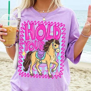 May include: A lilac-colored t-shirt featuring a cartoon horse with a saddle and the text "HOLD YOUR". The design is framed by a pink wavy border with stars. The shirt has short sleeves.