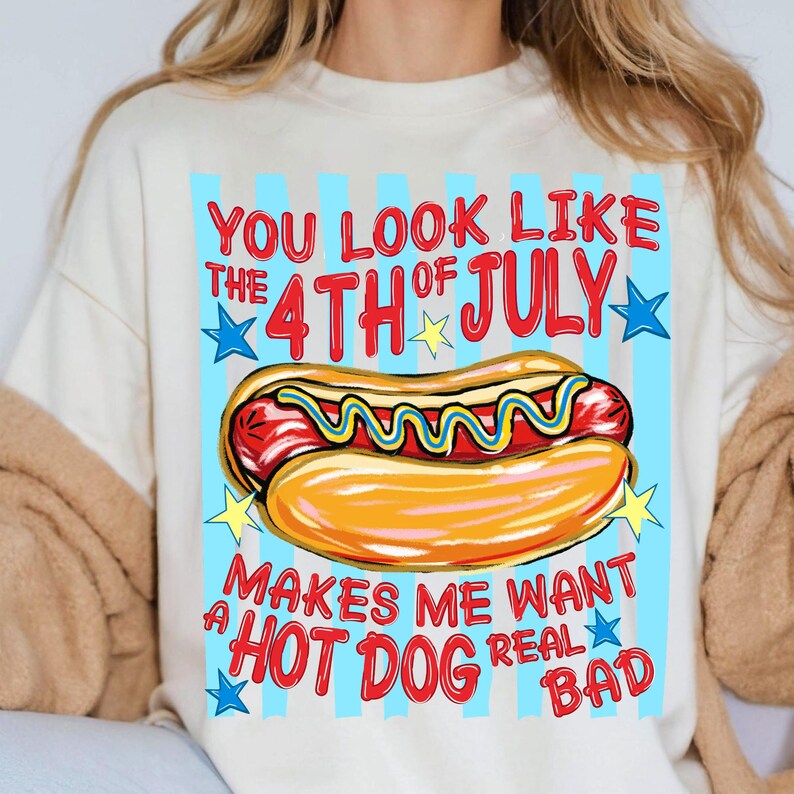 May include: Cream-colored t-shirt with a graphic of a hot dog and the text "You look like the 4th of July makes me want a hot dog real bad." The design includes red, yellow, and blue colors, with star accents and a blue striped background.