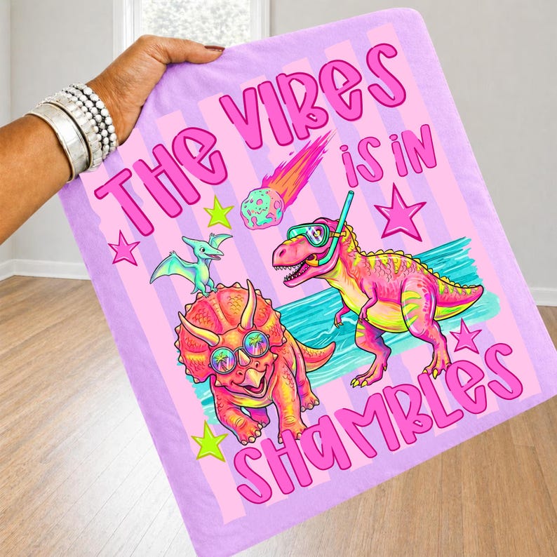 Peut inclure: Une couverture rectangulaire, violet clair, avec des rayures roses et le texte "THE VIBES IS IN SHAMBLES" en rose. La couverture pr&eacute;sente des dinosaures de dessins anim&eacute;s, des &eacute;toiles et une m&eacute;t&eacute;orite aux couleurs vives.