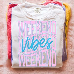 K&ouml;nnte beinhalten: Ein Stapel gefalteter T-Shirts in verschiedenen Farben, mit einem wei&szlig;en Shirt obenauf. Das wei&szlig;e Shirt tr&auml;gt den Text "WEEKEND VIBES WEEKEND" in Rosa und Blau. Die Shirts liegen auf einer rosa Oberfl&auml;che.