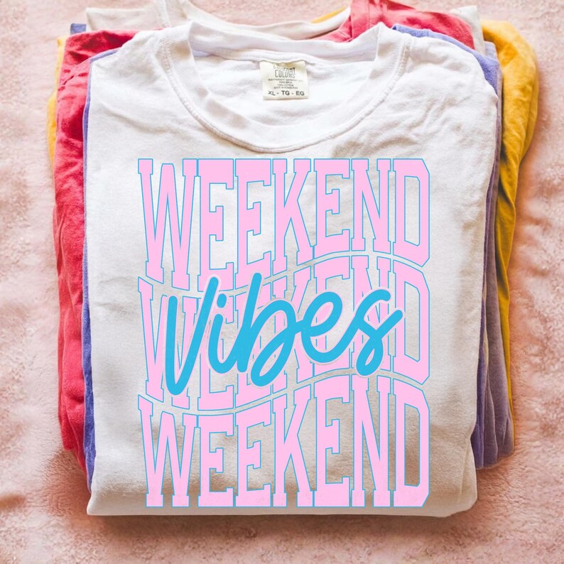 K&ouml;nnte beinhalten: Ein Stapel gefalteter T-Shirts in verschiedenen Farben, mit einem wei&szlig;en Shirt obenauf. Das wei&szlig;e Shirt tr&auml;gt den Text "WEEKEND VIBES WEEKEND" in Rosa und Blau. Die Shirts liegen auf einer rosa Oberfl&auml;che.