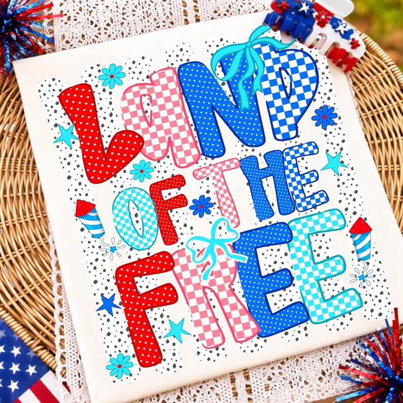 Puede incluir: Una camiseta blanca con el texto LAND OF THE FREE en letras grandes, coloridas y estampadas. El dise&ntilde;o incluye lunares y cuadros rojos, rosas y azules, acentuados con lazos azul claro, peque&ntilde;as flores, estrellas y gr&aacute;ficos de fuegos artificiales.