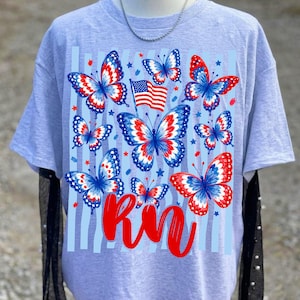 Peut inclure: Un t-shirt gris chin&eacute; avec une impression graphique de plusieurs papillons aux motifs d'ailes rouges, blancs et bleus. Le design comprend un petit drapeau am&eacute;ricain, des &eacute;toiles rouges et bleues, des rayures verticales bleu clair et le texte RN en rouge.