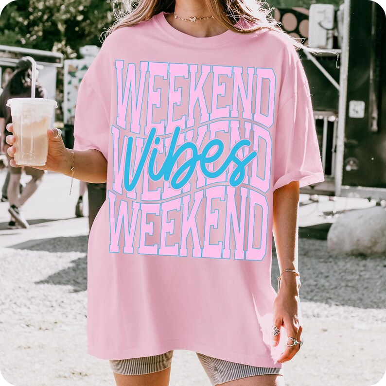 K&ouml;nnte beinhalten: Hellrosa T-Shirt mit dem Schriftzug "WEEKEND Vibes WEEKEND" in Blau und Hellrosa. Das Shirt hat eine lockere Passform und scheint aus einem weichen Material zu sein. Der Gesamtstil ist l&auml;ssig und bequem.