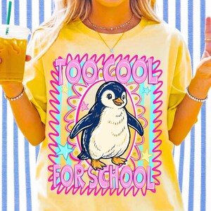 K&ouml;nnte beinhalten: Gelbes T-Shirt mit einem Pinguin-Cartoon-Motiv. Der Pinguin ist blau, wei&szlig; und orange, umgeben von einem rosa und blauen Design mit dem Text "TOO COOL FOR SCHOOL". Das Shirt hat kurze &Auml;rmel.