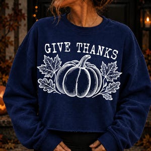 Puede incluir: Una sudadera azul marino de manga larga con un estampado gr&aacute;fico blanco de una calabaza entre hojas de arce y roble. El texto GIVE THANKS aparece en una fuente serif blanca sobre la ilustraci&oacute;n de la calabaza. Tiene cuello redondo.