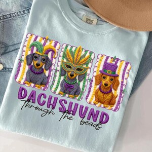 Puede incluir: Camiseta azul claro con un dise&ntilde;o bordado de tres perros salchicha con disfraces de Mardi Gras. El texto "DACHSHUND Through the beads" est&aacute; bordado debajo en morado.