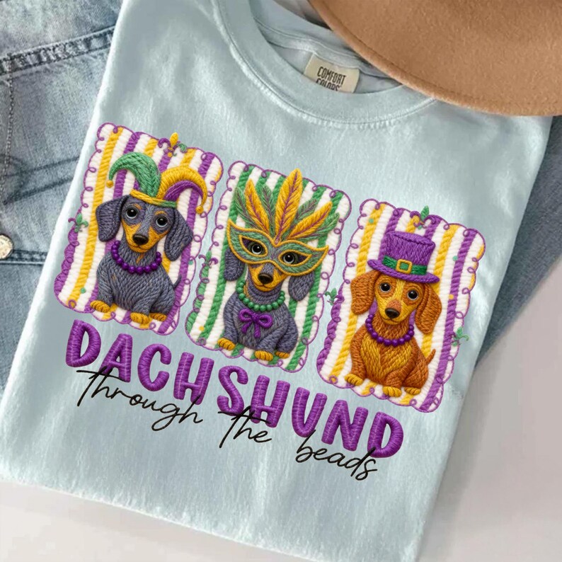 Puede incluir: Camiseta azul claro con un dise&ntilde;o bordado de tres perros salchicha con disfraces de Mardi Gras. El texto "DACHSHUND Through the beads" est&aacute; bordado debajo en morado.