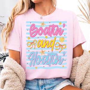 以下が含まれることがあります： 「Boatin' Sand & Floatin'」の文字が遊び心のあるフォントでプリントされたピンクのTシャツ。デザインには青いストライプ、ヒトデ、貝殻、星が含まれています。カジュアルな夏のトップス。