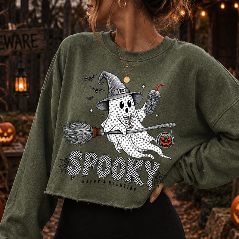 May include: An olive green cropped sweatshirt featuring a graphic of a white ghost wearing a witch hat, holding a drink, and riding a broomstick with a jack-o'-lantern. The text reads SPOOKY and HAPPY HAUNTING in a black and white chequered font.