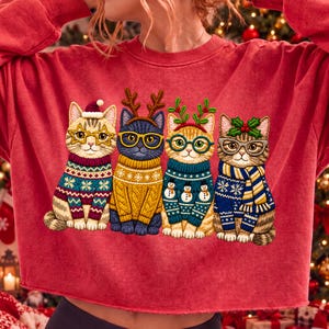 May include: A red long-sleeve sweatshirt featuring a graphic of four cats wearing holiday sweaters and glasses. The cats are adorned with festive accessories including a Santa hat, reindeer antlers, and holly leaves, set against a solid red background.