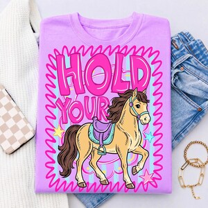 May include: A light purple t-shirt featuring a cartoon horse with a saddle. The text "HOLD YOUR" is in pink, outlined in a wavy pattern. The horse is tan with a brown mane and tail, and the background has stars and stripes.