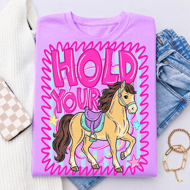 May include: A light purple t-shirt featuring a cartoon horse with a saddle. The text "HOLD YOUR" is in pink, outlined in a wavy pattern. The horse is tan with a brown mane and tail, and the background has stars and stripes.