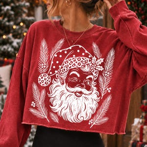 Puede incluir: Una sudadera roja de manga larga y corte corto con un estampado gr&aacute;fico blanco de un Santa Claus gui&ntilde;ando un ojo, con un gorro de lunares y estampado de leopardo, rodeado de ramas de pino y bayas de acebo estilizadas en el centro.