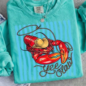 Op de afbeelding: Een turquoise Comfort Colors sweatshirt met een afbeelding van een rode kreeft met een gele cowboyhoed en een touwlasso. De woorden "Yee Claw" zijn geschreven in een lettertype in touwstijl. Verticale blauwe strepen staan achter de kreeft.