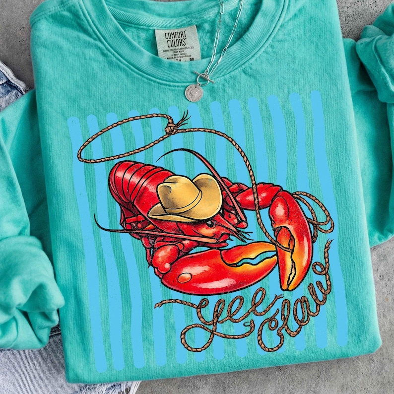 Op de afbeelding: Een turquoise Comfort Colors sweatshirt met een afbeelding van een rode kreeft met een gele cowboyhoed en een touwlasso. De woorden "Yee Claw" zijn geschreven in een lettertype in touwstijl. Verticale blauwe strepen staan achter de kreeft.