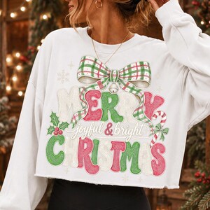 K&ouml;nnte beinhalten: Ein wei&szlig;es, lang&auml;rmeliges, bauchfreies Sweatshirt mit einem Grafikdruck einer gro&szlig;en gr&uuml;n-roten karierten Schleife &uuml;ber dem Text Merry Christmas in einer stilisierten Schriftart, erg&auml;nzt durch den Schriftzug joyful & bright.