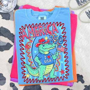 Puede incluir: Camiseta azul claro con un gr&aacute;fico de un caim&aacute;n con sombrero de vaquero y una camiseta "I ❤️ USA", sosteniendo una bandera americana. La camiseta tiene el texto "AMERICA 250" en rojo y azul. La camiseta est&aacute; doblada sobre otras camisetas de colores.