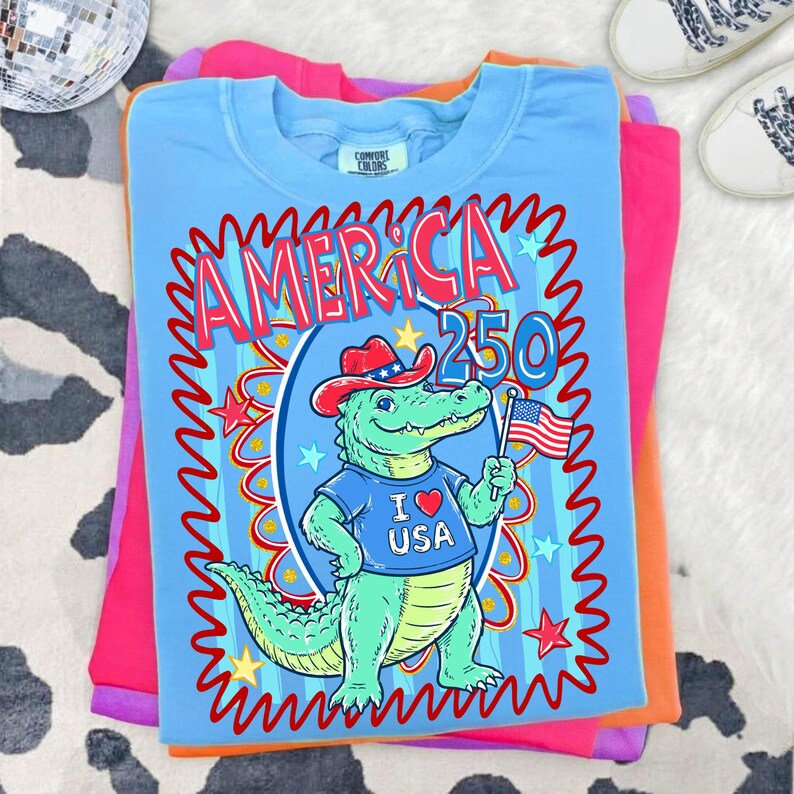 Puede incluir: Camiseta azul claro con un gr&aacute;fico de un caim&aacute;n con sombrero de vaquero y una camiseta "I ❤️ USA", sosteniendo una bandera americana. La camiseta tiene el texto "AMERICA 250" en rojo y azul. La camiseta est&aacute; doblada sobre otras camisetas de colores.