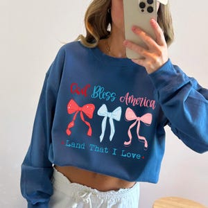 Może przedstawiać: Granatowa bluza z napisem "God Bless America" i "Land That I Love" w kolorze czerwonym, jasnoniebieskim i r&oacute;żowym. Projekt zawiera trzy grafiki kokardek w kolorze czerwonym, jasnoniebieskim i r&oacute;żowym.