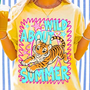 K&ouml;nnte beinhalten: Gelbes T-Shirt mit einem Tiger-Grafik und dem Text "WILD ABOUT SUMMER" in T&uuml;rkis und Pink. Der Tiger ist orange und schwarz, umgeben von Sternen und einem wellenf&ouml;rmigen rosa Rahmen.