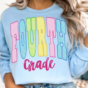 May include: A light blue crewneck sweatshirt featuring the text "FOURTH" in large, multicolored, vertically striped block letters, with the word "Grade" written below in a vibrant pink cursive script font across the chest area.