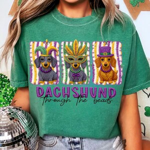 Puede incluir: Camiseta verde con un dise&ntilde;o de tres perros salchicha con disfraces de Mardi Gras. Cada perro lleva un accesorio diferente: un gorro de buf&oacute;n, una m&aacute;scara de plumas y un sombrero de copa. El texto "DACHSHUND through the beads" est&aacute; debajo.