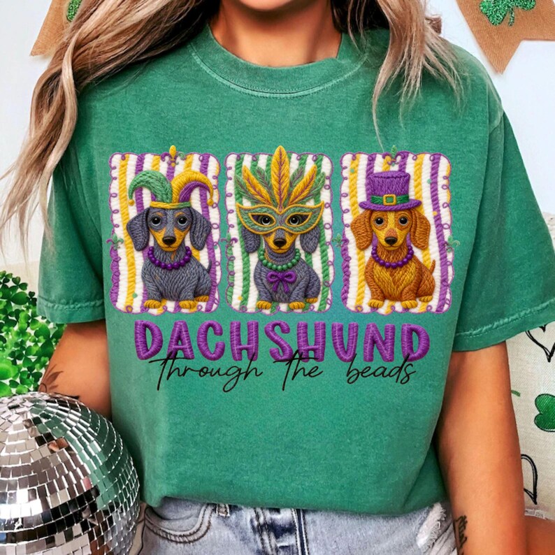 Puede incluir: Camiseta verde con un dise&ntilde;o de tres perros salchicha con disfraces de Mardi Gras. Cada perro lleva un accesorio diferente: un gorro de buf&oacute;n, una m&aacute;scara de plumas y un sombrero de copa. El texto "DACHSHUND through the beads" est&aacute; debajo.
