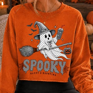May include: An orange long-sleeve sweatshirt featuring a graphic of a ghost wearing a witch hat, holding a drink, and riding a broomstick with a jack-o'-lantern. The design includes bats, stars, and the text "SPOOKY HAPPY HAUNTING" in a checkered font.