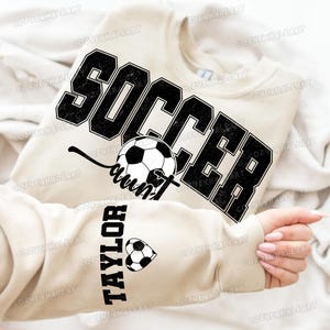 May include: Beige sweatshirt with the word "SOCCER" in large, distressed black letters. Below the word "SOCCER" is the word "aunt" with a soccer ball graphic. The sleeve has the name "TAYLOR" and a soccer ball heart.