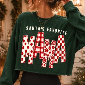 K&ouml;nnte beinhalten: Ein dunkelgr&uuml;nes, lang&auml;rmeliges, bauchfreies Sweatshirt mit dem wei&szlig;en Schriftzug SANTA'S FAVORITE &uuml;ber dem Wort MAMA. Die Buchstaben von MAMA sind mit rot-wei&szlig;en Mustern gef&uuml;llt, darunter Punkte, Karos, Blumenmotive und ein Schachbrettmuster.