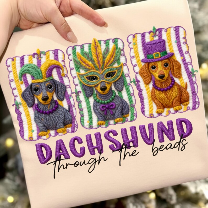 Puede incluir: Camiseta beige claro con tres perros salchicha bordados con disfraces de Mardi Gras. Los perros llevan sombreros, m&aacute;scaras y collares festivos. El texto "DACHSHUND Through the beads" est&aacute; bordado en morado.