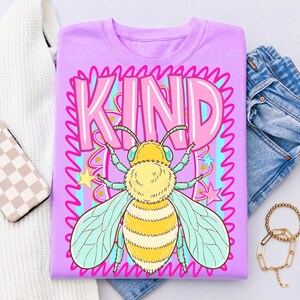 May include: A lilac-colored t-shirt featuring a large graphic of a bee with yellow and white stripes, blue wings, and the word "KIND" in pink. The design includes stars and wavy lines.
