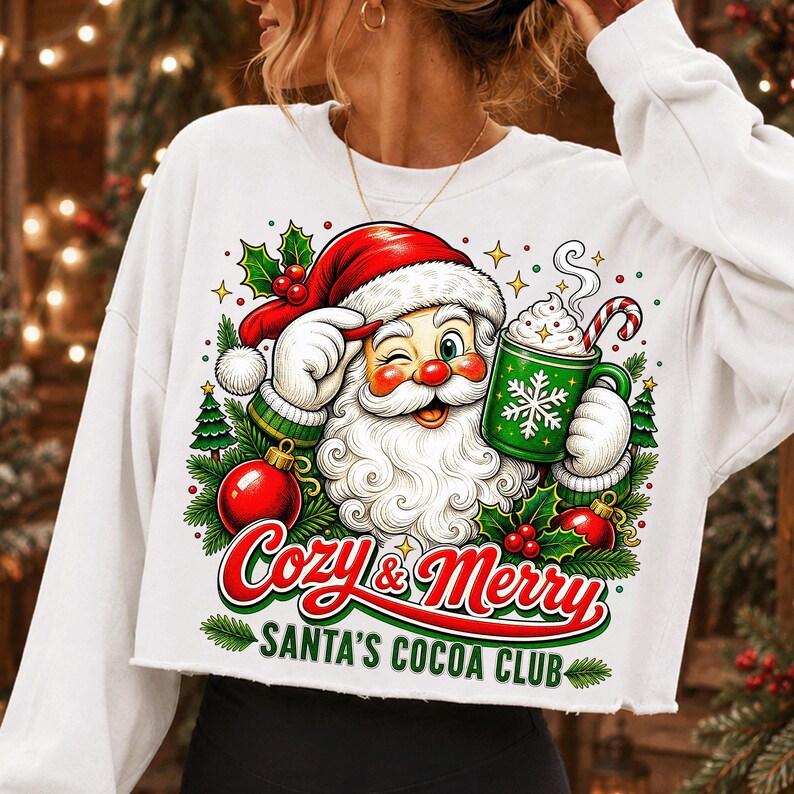 K&ouml;nnte beinhalten: Ein wei&szlig;es Langarm-Sweatshirt mit einer Grafik eines zwinkernden Weihnachtsmanns, der eine gr&uuml;ne Tasse mit Schneeflockenmuster und eine Zuckerstange h&auml;lt. Der Text lautet Cozy & Merry SANTA'S COCOA CLUB, umgeben von Stechpalmen und Tannenb&auml;umen.