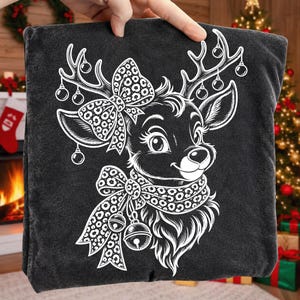 May include: A dark grey t-shirt featuring a white line art graphic of a reindeer head. The reindeer has a large leopard print bow between its antlers, which are decorated with hanging ornaments. A leopard print scarf with two jingle bells is worn.