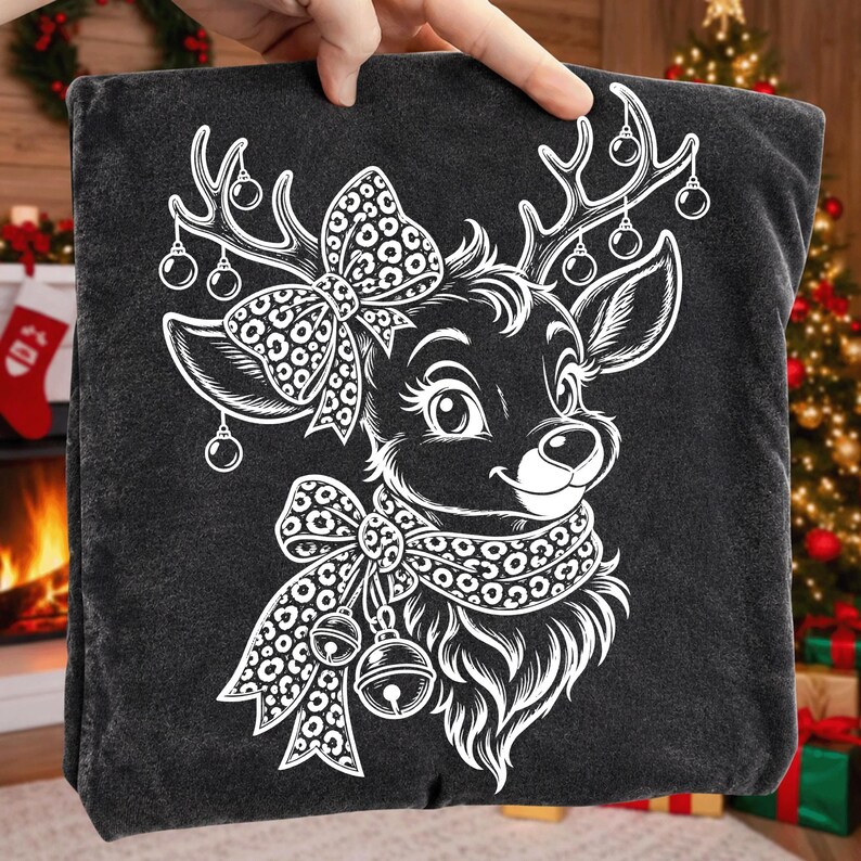 May include: A dark grey t-shirt featuring a white line art graphic of a reindeer head. The reindeer has a large leopard print bow between its antlers, which are decorated with hanging ornaments. A leopard print scarf with two jingle bells is worn.
