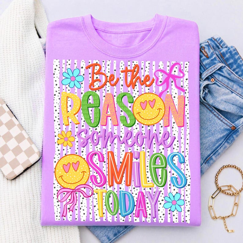 May include: A lilac-colored t-shirt with the text "Be the REASON someone SMILES TODAY" in colorful, playful lettering. The design includes smiley faces, flowers, and bows on a striped background.