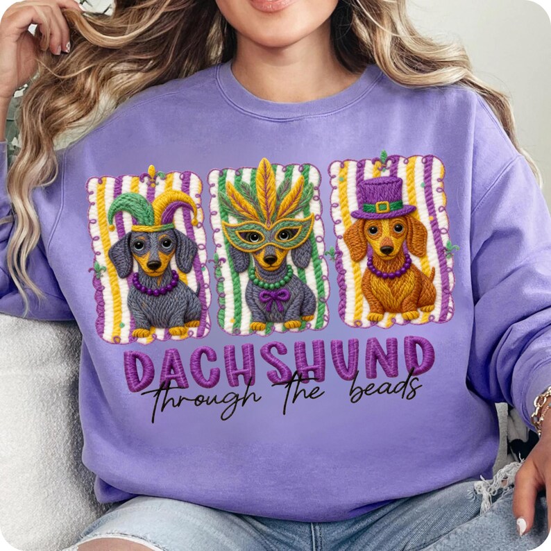 Puede incluir: Una sudadera lila con un dise&ntilde;o de tres perros salchicha con disfraces de Mardi Gras. Cada perro est&aacute; en un dise&ntilde;o enmarcado con rayas verticales y un borde decorativo. El texto "DACHSHUND Through the beads" est&aacute; impreso debajo.