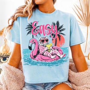 K&ouml;nnte beinhalten: Hellblaues T-Shirt mit einem Tiger, der eine Herzsonnenbrille tr&auml;gt und sich auf einem rosa Flamingo-Schwimmer entspannt, einen Cocktail h&auml;lt. Das Wort "Easy" steht in Pink &uuml;ber dem Tiger. Palmen und ein Sonnenuntergang sind im Hintergrund.