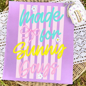 K&ouml;nnte beinhalten: Eine lavendelfarbene Tragetasche mit vertikalen rosa und wei&szlig;en Streifen und den Worten "Made for Sunny Days" in T&uuml;rkis, Rosa und Gelb. Eine kleine, blumenmusternde Handtasche mit goldener Kette ist oben rechts zu sehen.